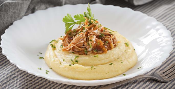 Fotografia em tons de cinza, branco e amarelo, prato branco ao centro com polenta, frango desfiado e salsa, ao lado um garfo base, sobre toalha com listras brancas e cinza e ao fundo guardanapo cinza.