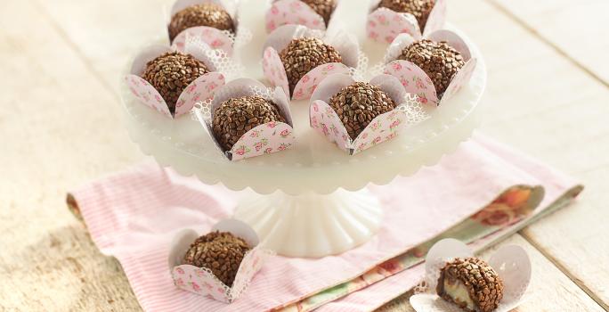 fotografia em tons de cinza, rosa e marrom de uma bancada cinza vista de cima, contém um suporte branco para servir sobremesas com forminhas rosas com brigadeiros dentro, e abaixo um pano rosa com 2 forminhas e ambas contém brigadeiro