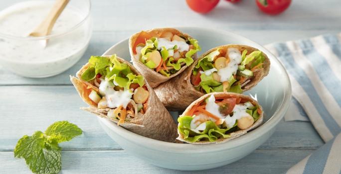 Foto em tons coloridos da receita de wrap vegetariano com molho de hortelã servida em 4 porções dentro de um bowl de porcelana branca sobre uma mesa de madeira azul clara com um paninho listrado azul e um pote de iogurte natural