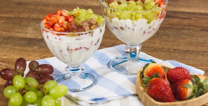 Fotografia em tons de azul e bege em uma bancada e madeira escura, um pano listrado em azul e bege, duas taças de vidro com a sobremesa de creme com morangos e uvas picados. Ao lado, um potinho com morangos em cima e cachos de uvas.