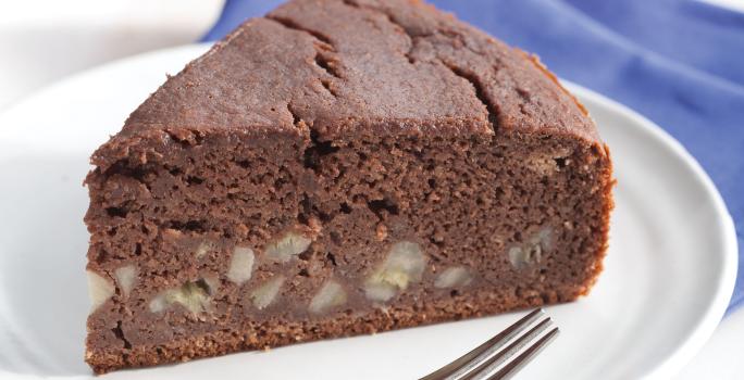 Fotografia em tons de azul em uma mesa de madeira branca com um pano azul, um prato branco raso com uma fatia de bolo de chocolate com banana.