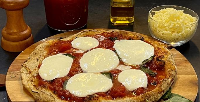 Foto da receita de pizza napolitana servida sobre uma tábua de madeira redonda com folhas manjericão ao lado. Ao fundo, há um bowl de vidro com muçarela, um vidro de azeite, um moedor de sal e um vidro de molho pomodoro