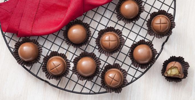 Fotografia em tons de chocolate, branco e vermelho, ao centro vários bombons de chocolate em forminhas de papel marrom sobre grelha redonda, ao lado guardanapo vermelho e bombom aberto, tudo sobre bancada de madeira clara.