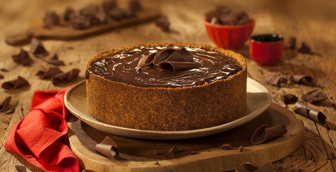 Fotografia em tons de marrom em uma bancada de madeira de cor marrom. Ao centro, uma tábua de madeira contendo o cheesecake e ao redor há pedaços de chocolates espalhados.