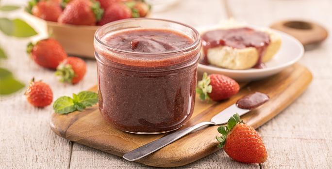 Foto em tons de vermelho da receita de geleia cremosa de morango servida em um recipiente de vidro com uma faca arredonda prateada ao lado e morangos decorativos. Ao fundo há um prato com um pão e a geleia por cima, além de uma cesta com morangos