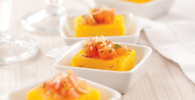 Fotografia em tons de laranja em uma bancada de madeira clara, um pano bege, quatro potinhos brancos pequenos com a polenta com tomate dentro de cada um.