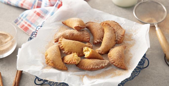 Foto da Receita de Pastel de Grão-de-bico. Observa-se um prato com papel absorvente e os pasteizinhos em cima polvilhados de canela e açúcar. Ao lado esquerdo, dois paus de canela e, ao direito, uma peneira com.