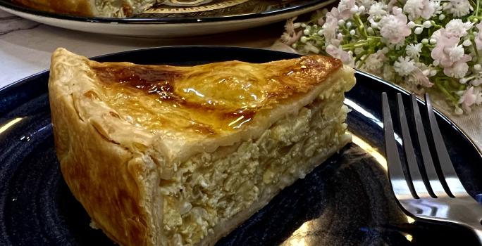 Foto da receita de Torta de Cebola e Ovos. Observa-se um pedaço grande de torta em um prato preto com decoração de flores brancas.