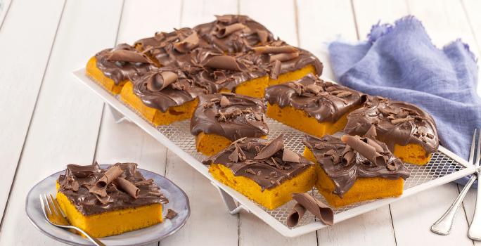 Fotografia em tons de azul em uma bancada de madeira branca, um pano azul, um apoiador de grelha branco com vários pedaços do bolo de cenoura com cobertura de chocolate. Ao lado, um prato pequeno com uma fatia do bolo.