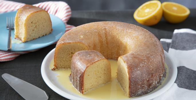 Fotografia em tons de amarelo em uma bancada de madeira cor preta. Ao centro, um prato branco contendo o bolo com uma espátula ao lado. Ao fundo, há um pires azul com uma fatia do bolo e uma laranja partida ao meio.