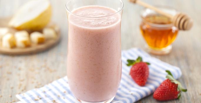 Fotografia em tons de rosa em uma bancada de madeira clara, um paninho listrado azul, um copo de vidro alto com a vitamina de morango. Ao lado, dois morangos. Ao fundo, um potinho com mel e um pratinho com pedaços pequenos de pera.