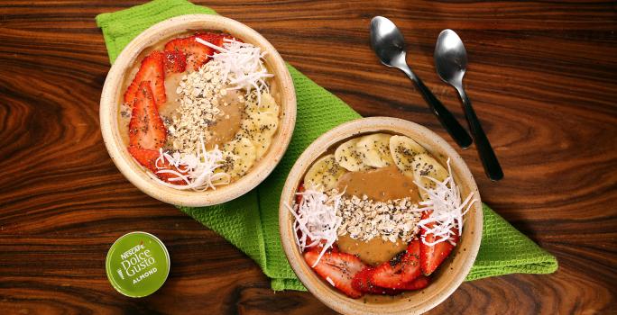 Fotografia em tons de verde em uma bancada de madeira escura, um pano verde, dois bowls bege com morango, banana e lascas de coco e duas colheres ao lado.