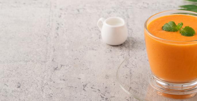 Fotografia em tons claros de uma vitamina laranja com folhas de hortelã por cima em um copo de vidro, apoiado sobre um prato pequeno de vidro. Ao fundo, um pote pequeno branco de vidro, sobre uma mesa em tons de cinza.