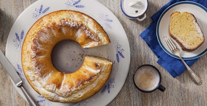 Fotografia em tons de cinza, branco e azul de uma bancada cinza e um prato banco com detalhes azuis, sobre ele o bolo de milho. Ao lado duas xícaras de café com leite e um paninho azul com um prato branco de bordas azuis com uma fatia do bolo e um garfo.