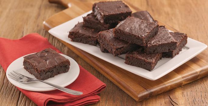 Fotografia em tons de marrom e vermelho de uma bancada de madeira com uma tábua de madeira, sobre ele um papel manteiga com pedaços de brownie. Ao  fundo um prato com um pedaço de brownie e um garfo.