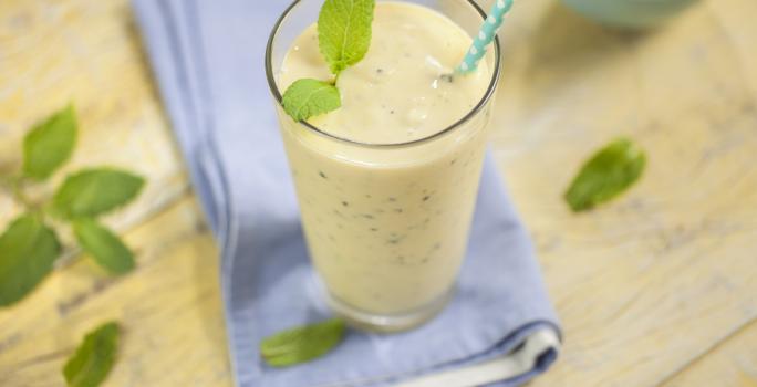 Fotografia em tons de marrom e azul de uma bancada de madeira com um paninho azul, sobre ele um copo com smoothie com um canudo. Ao lado folhinhas de hortelã e um recipiente redondo com hortelã.