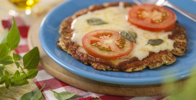 fotografia em tons de azul, vermelho e marrom de uma bancada marrom vista de cima, contém uma tábua redonda, um prato redondo azul com uma mini pizza coberta com queijo, duas fatias de tomate e manjericão e ao lado um garfo para servir.