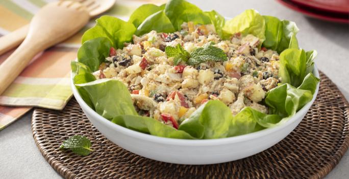 Fotografia em tons de verde em uma bancada de madeira, um pano com listras coloridas, um jogo americano redondo de vime escuro e um recipiente branco redondo fundo com a salada marroquina com vários legumes dentro dela.