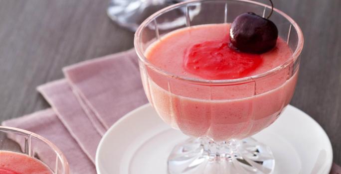 Fotografia em tons de vermelho em uma mesa de madeira com um pano rosa escuro e um prato pequeno branco apoiado nele. Em cima do prato, uma taça de vidro com a mousse de cereja e uma cereja por cima para enfeita. Ao lado e ao fundo, mais duas taças.