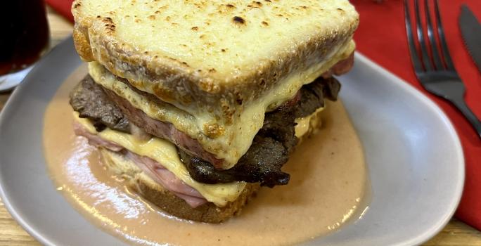 Foto da receita de Francesinha. Oberva0se um prato de cerâmica cinza com o sanduíche por cima, com queijo gratinado e um molho amarronzado.