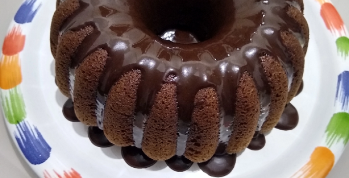 Foto em tons de marrom da receita de bolo de café servida com uma cobertura de chocolate por cima e sobre um prato branco com a borda colorida