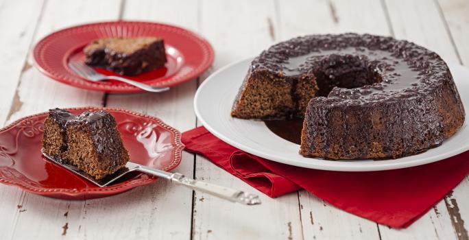 Fotografia em tons de vermelho em uma bancada de madeira clara com um prato branco ao centro com o bolo de chocolate cortado ao meio e um paninho vermelho embaixo. Ao lado, dois pratos vermelhos com uma fatia de bolo em cada um.