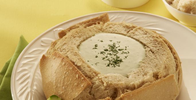 Fotografia em tons de amarelo, branco e bege, com prato branco contendo pão italiano recheado com creme, dois espetos com legumes e um guardanapo verde ao lado, ao fundo potinhos com legumes, espetinhos e pão, tudo sobre toalha amarela.