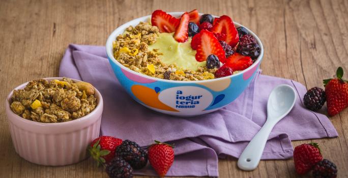 Fotografia em tons de lilás em uma bancada de madeira clara, um bowl colorido com o iogurte de abacate com frutas vermelhas e granola decorando. Ao lado, um potinho com granola e frutas espalhadas na bancada.