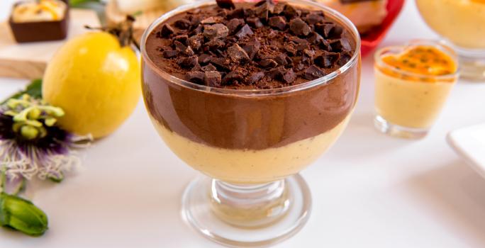 Fotografia de uma sobremesa de maracujá, chocolate e biscoito em uma taça, que está sobre uma mesa branca. Ao fundo, diversas opções de mousse de maracujá, a fruta inteira e uma tábua de madeira clara.