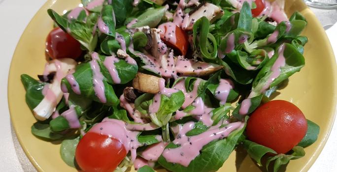Fotografia vista de cima de uma salada de alface, cenoura, tomates cereja, pepino e beterraba com molho à base de iogurte e ketchup. A salada está em um prato amarelo que está sobre uma mesa branca.