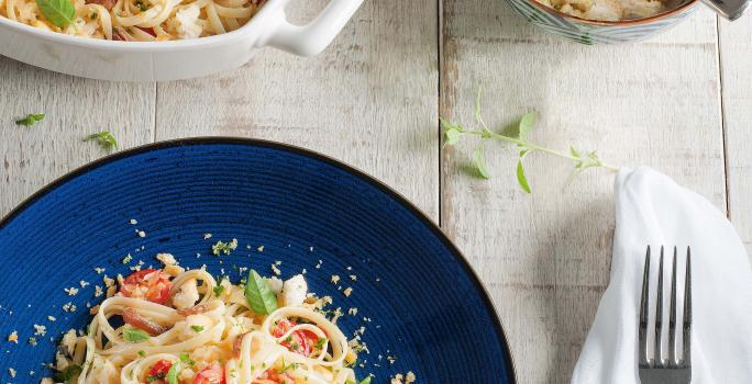 Fotografia em tons de azul em uma mesa de madeira clara com um guardanapo branco, um prato azul redondo com o macarrão com tomates e anchova.