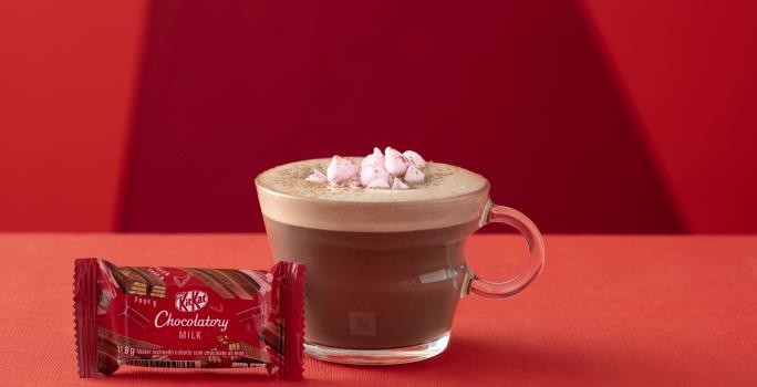 Foto da receita de Chocolate Quente. Observa-se um fundo vermelho com o chocolate quente em uma xícara de vidro transparente. Na frente, um kit kat fechado ao leite.