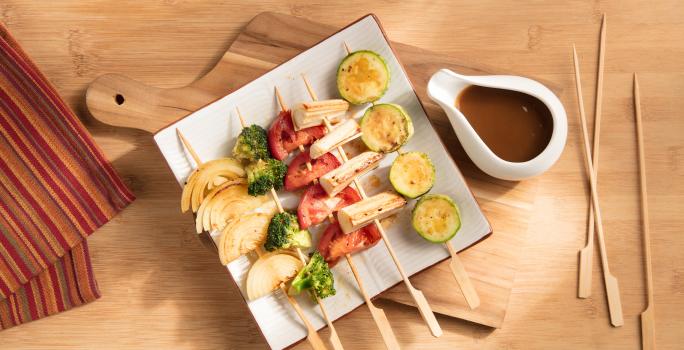 Fotografia em tons de laranja em uma mesa de madeira, uma tábua de madeira, um pano listrado com cores alaranjada e um prato quadrado com vários espetinhos de legumes. Ao lado, um potinho com o molho shoyu.