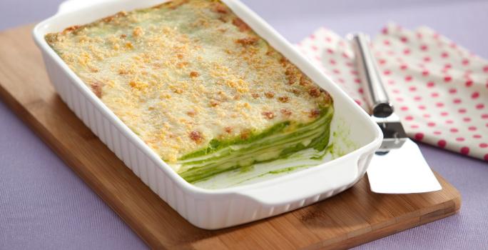 Fotografia em tons de branco e verde, com refratário branco contendo uma lasanha de espinafre gratinada, sobre uma tábua de cozinha com uma espátula de servir sobre um guardanapo branco com bolinhas vermelhas em uma bancada lilás.