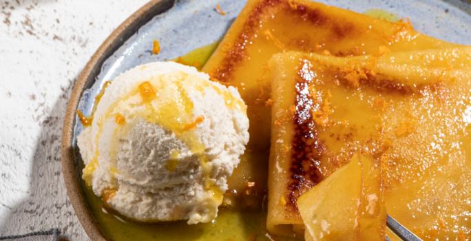 Foto de um prato em tom claro com a receita de Crepe Suzette, servido com calda amarela e uma bola de sorvete. O prato está em uma bancada clara, com. um pote de calda amarela e um guardanapo ao lado.