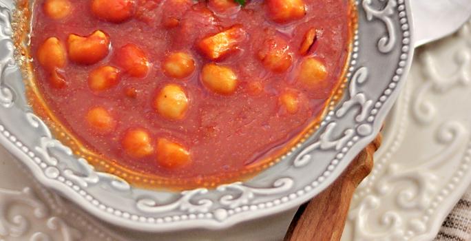 Fotografia vista de cima de uma sopa de tomates com nhoquettes em um prato fundo desenhado em tons de cinza, apoiado sobre um prato raso branco desenhado com uma colher apoiada. Os pratos estão sobre uma toalha de mesa marrom.