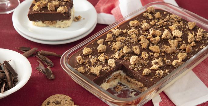 Fotografia de um pavê de chocolate com biscoito em uma travessa retangular transparente, com pedaços de biscoitos polvilhados por cima. Ao fundo uma toalha de mesa vermelha, com um prato de sobremesa branco servind um pedaço do pavê.