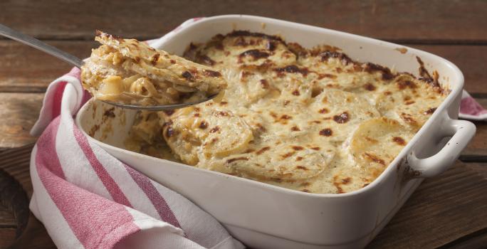 Fotografia em tons de marrom, branco e rosa de uma bancada marrom vista de frente. Contém um recipiente quadrado branco com alças e batatas e queijo gratinado, por cima uma colher com um pedaço da receita e ao lado um pano listrado nas cores rosa e branco