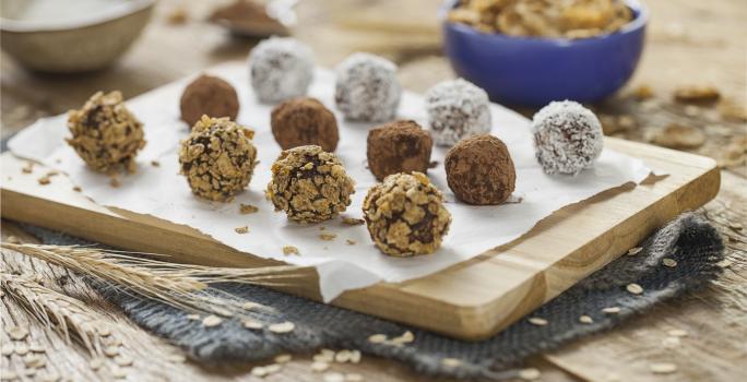 fotografia em tons de azul em uma bancada de madeira clara, uma tábua de madeira, um paninho azul escuro e vários docinhos parecidos com brigadeiros, mas feitos com Nesfit e sem açúcar. Ao fundo, potinhos com Nesfit Cereais.