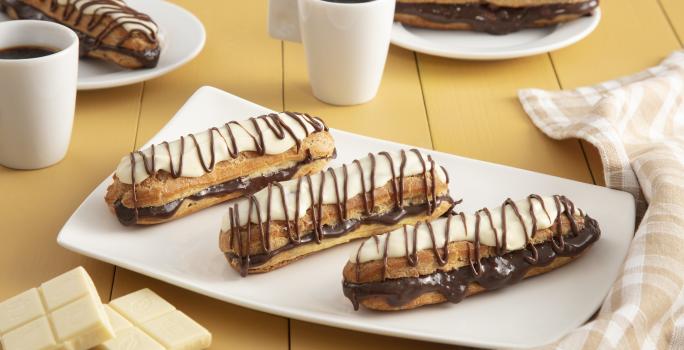 Fotografia em tons de amarelo em uma bancada de madeira com um pano xadrez brege, um recipiente retangular branco ao centro com três bombas de chocolate com cobertura de chocolate branco. Ao fundo, duas xícaras com café e dois pratinhos com duas bombas.