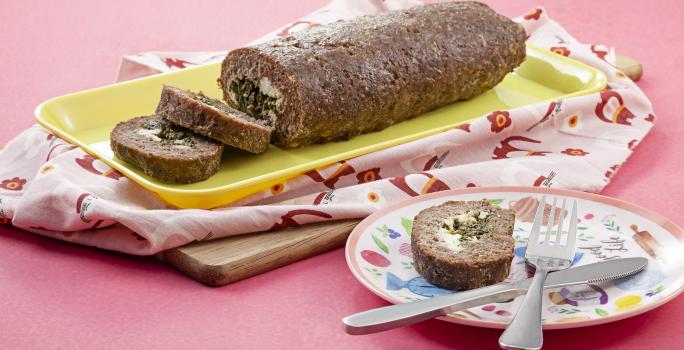 Fotografia em tons de rosa em uma mesa rosa com um pano rosa desenhado e um recipiente retangular amarelo com o bolo de carne com espinafre em cima dele. Ao lado, um prato colorido com uma fatia do bolo e um garfo e uma faca apoiados sobre ele.