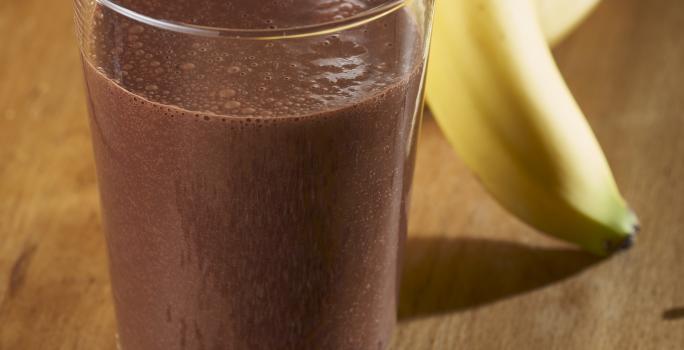Fotografia de um copo de vidro com uma vitamina de banana e chocolate Nescau. Ao fundo, um cacho de bananas, e na frente do copo, raspas de chocolate sobre uma mesa de madeira.