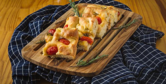 Foto aproximada de uma bancada com um tecido azul quadriculado sobre e nele uma tábua de madeira com pedaços de Focaccia, decorada com tomates-cereja e ramos de alecrim