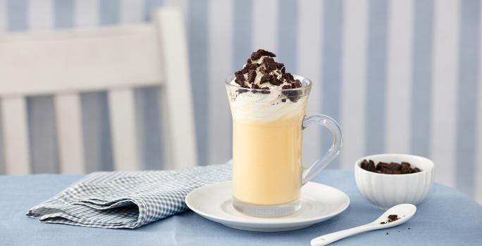 fotografia em tons de azul e branco de uma bancada azul vista de frente, contém um pratinho redondo branco com um copo transparente com a bebida dentro e por cima chantilly e biscoitos triturados para decorar. Ao lado um pano azul e do outro uma colher.