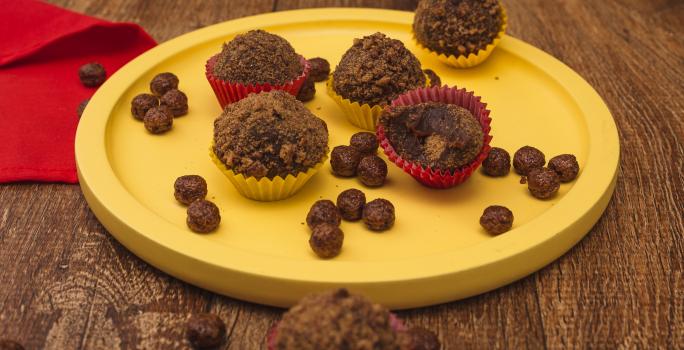 Fotografia em tons de amarelo em uma bancada de madeira escura, um pano vermelho, um prato amarelo redondo raso com brigadeiros de Nescau e recheados com Nescau Cereal.