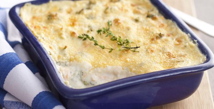 Fotografia em tons de azul em uma bancada de madeira clara, um pano branco com listras azuis, uma tábua de madeira, um recipiente quadrado azul com os legumes gratinados dentro dele.