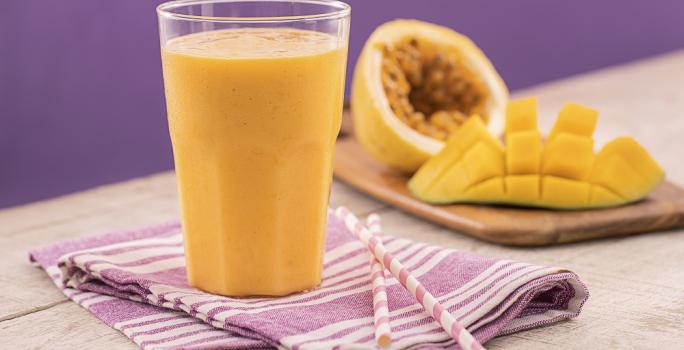 Foto em tons de roxo e amarelo da receita de smoothie de manga e maracujá servida em um copo de vidro largo sobre um pano lilás listrado com dois canudos ao lado. Ao fundo uma tábua de madeira com maracujá e manga cortados ao meio.