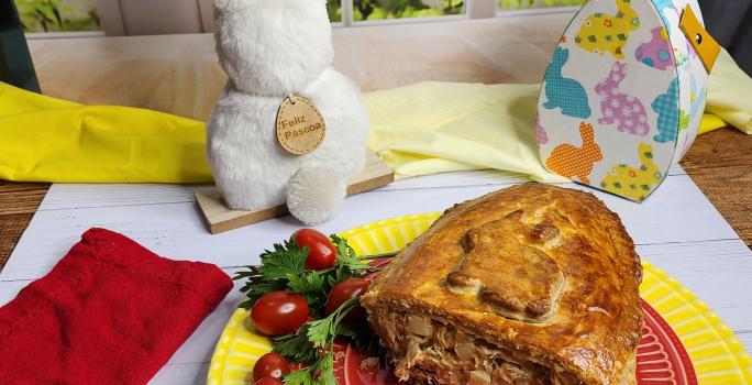 Foto da receita de Ovo de Colher Torta de Frango, servido cortado em um prato vermelho, sobre outro prato amarelo, em uma bancada branca decorada com itens de páscoa, como um coelho branco e uma caixinha em formato de ovo de páscoa