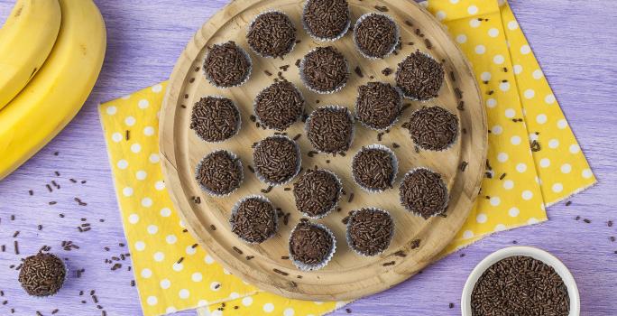Fotografia em tons de marrom, amarelo e lilás, com bancada lilás vista de cima, ao centro peça de madeira com brigadeiros sobre guardanapo amarelo com bolinhas brancas, ao lado potinho com granulado, um brigadeiro e e duas bananas.