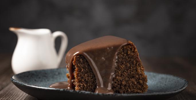 fotografia em tons de preto e marrom tirada de uma fatia de bolo de chocolate com calda por cima, e ao fundo um jarra branca